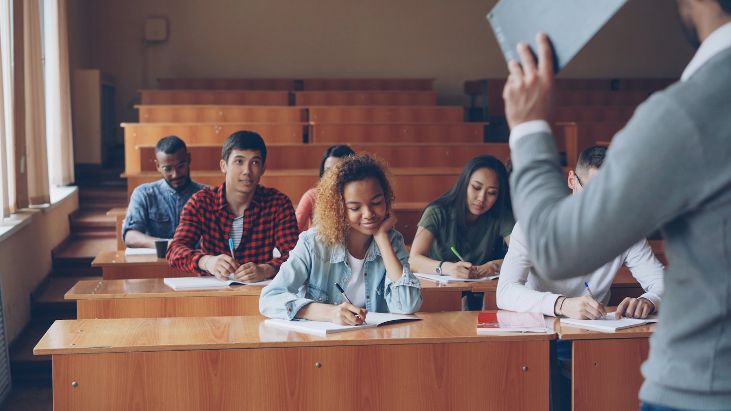 Studenti ascoltano attentamente un docente in una luminosa aula magna.